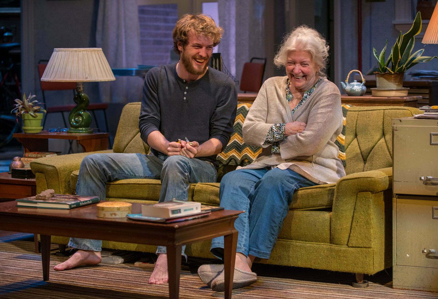 Two actors laughing together on the sofa in the living-room set of 4000 Miles