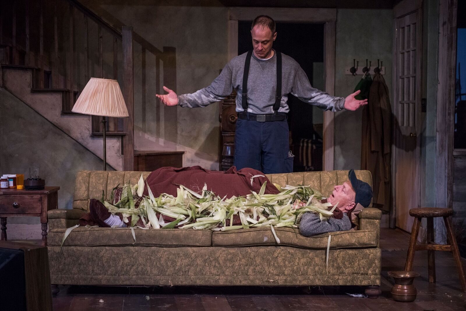 A figure lying on the sofa under a pile of corn husks while another stands over him