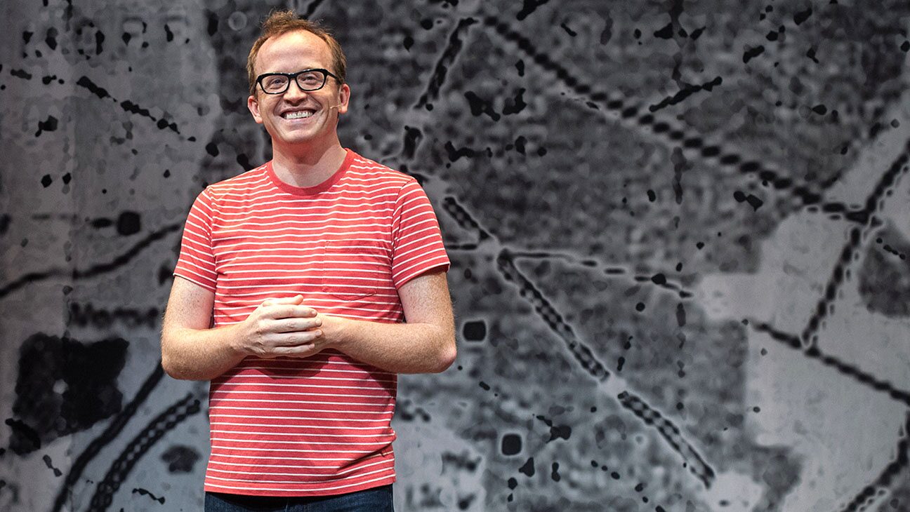 Chris Gethard smiling on stage in a red striped shirt in front of a projected map