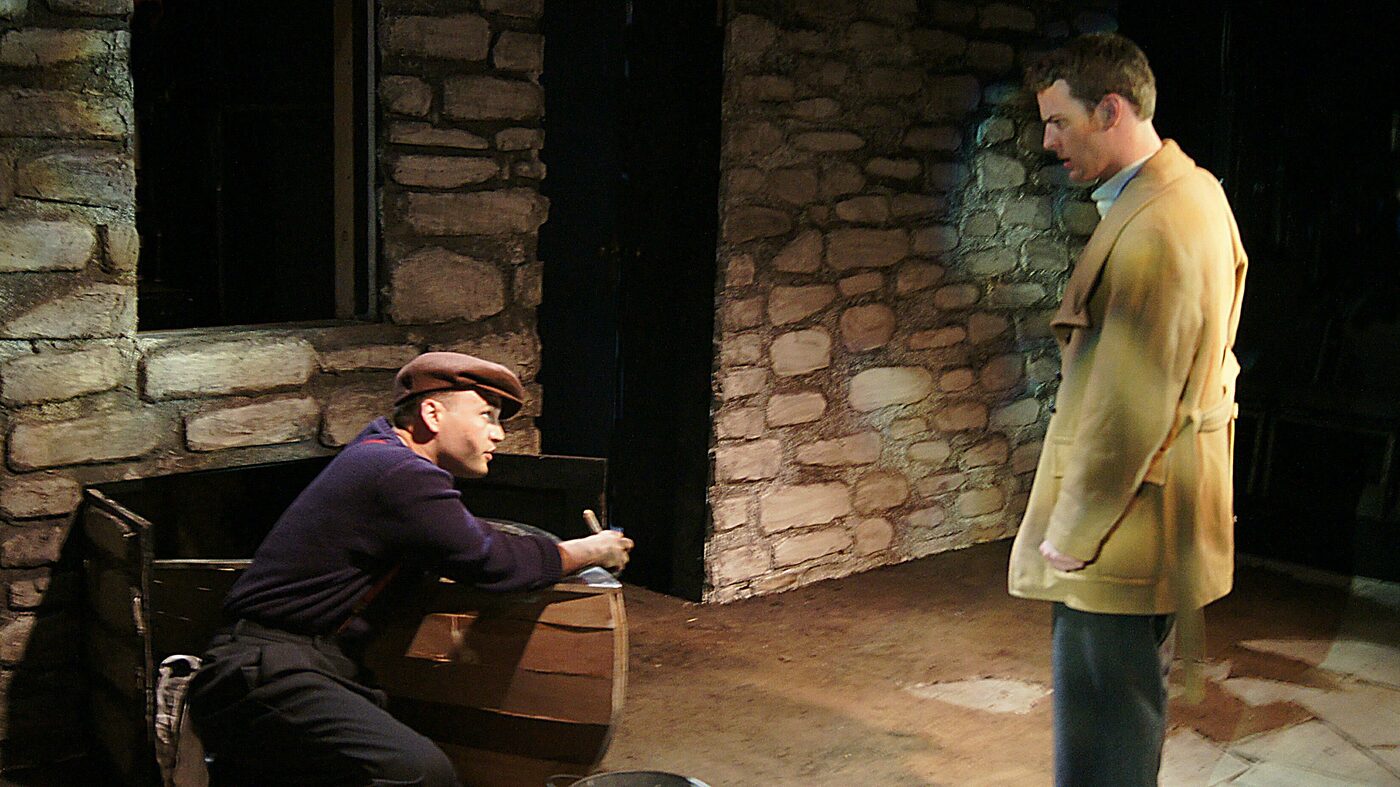 A man kneeling by a barrel and writing as another stands in a yellow coat