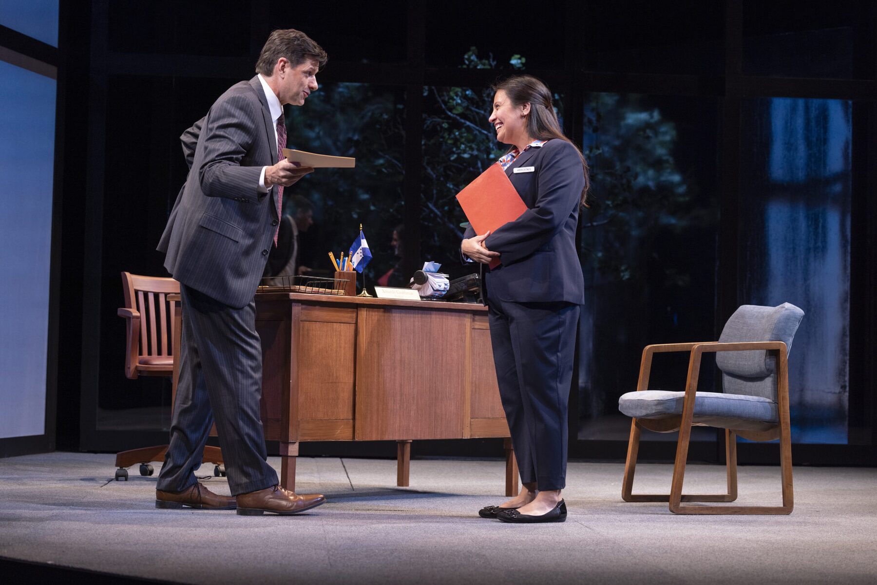 A man in a suit hands a folder across a desk to a woman in a uniform