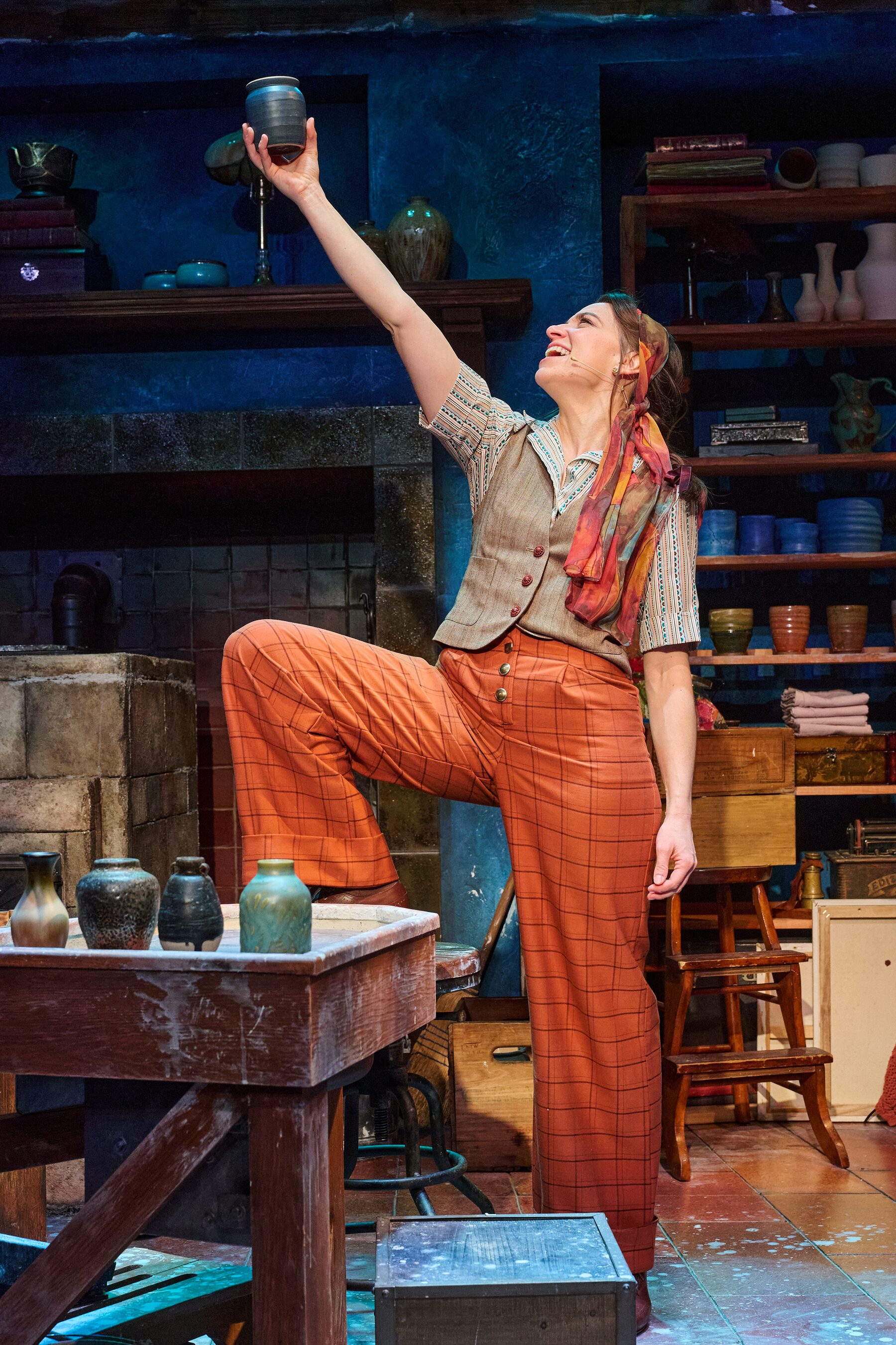 The potter in orange trousers stretching up to place a vase on a studio shelf