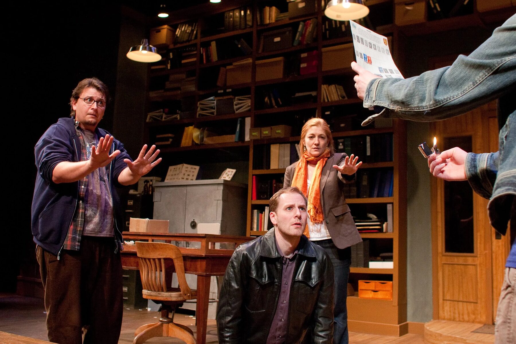 Four actors gather around a lit stamp in the bookshelf-lined set of Mauritius at TheatreSquared