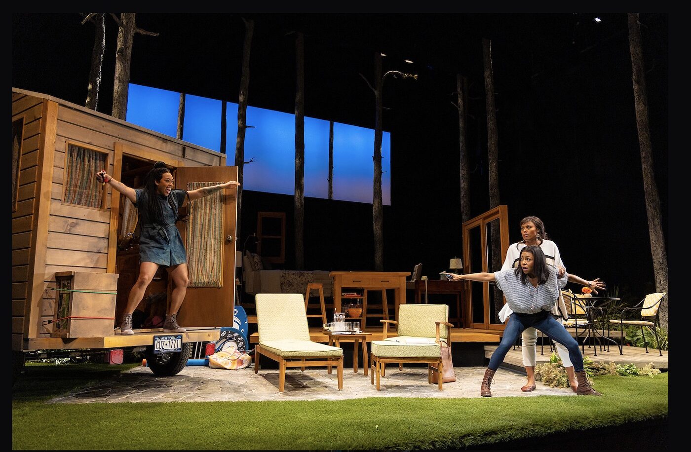 Actors on the lawn-and-trailer set of Our Daughters, Like Pillars, one in the trailer doorway with arms outstretched
