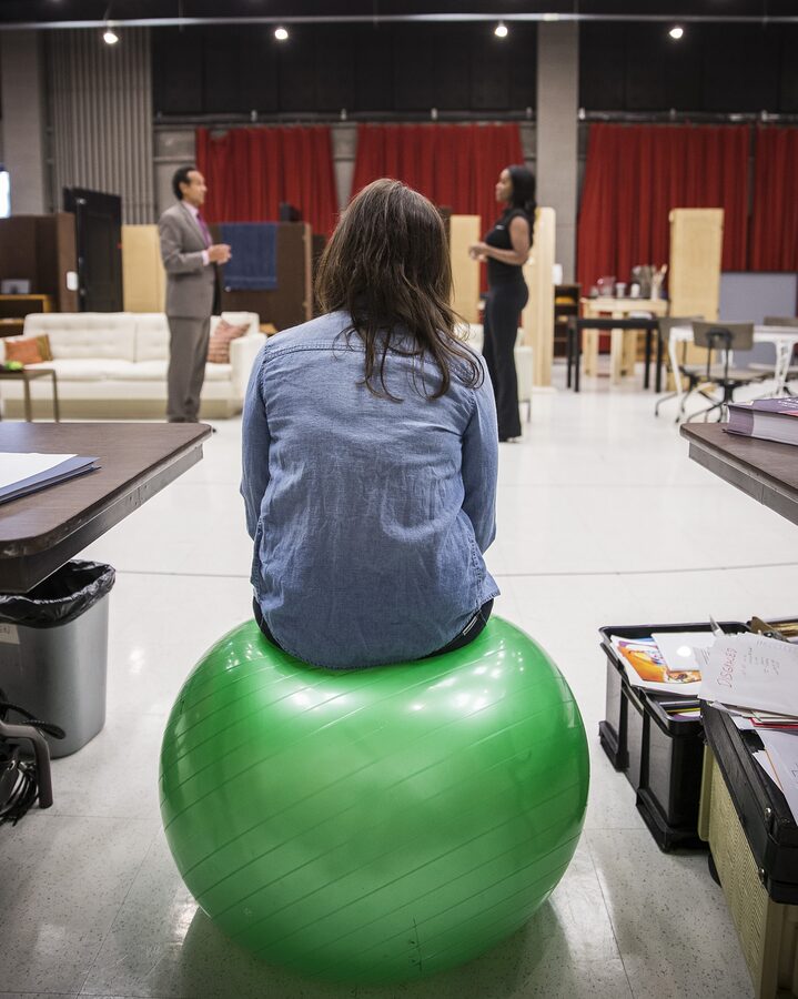 Kimberly Senior seated on an exercise ball, watching two actors rehearse a scene from Disgraced