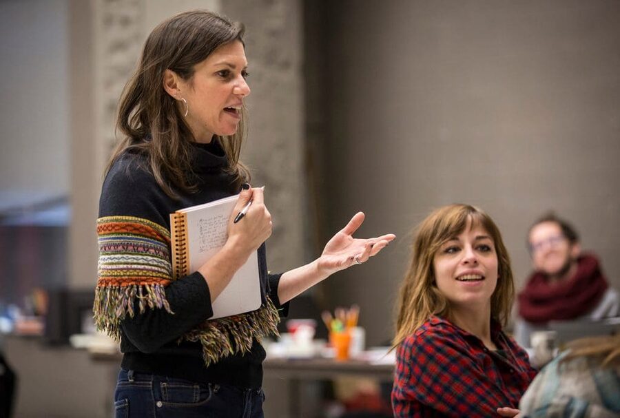 Kimberly Senior in a fringed sweater, notebook in hand, talking a scene through with actors seated at the rehearsal table