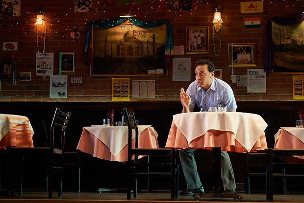 Aasif Mandvi seated alone at a table in the brick-walled restaurant set of Sakina&rsquo;s Restaurant