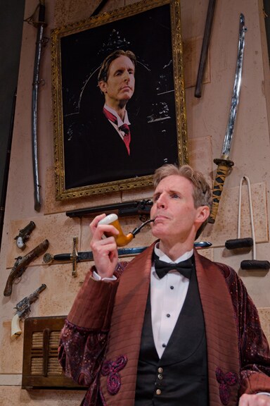 An actor in a velvet smoking jacket holding a pipe, in front of a wall of swords
