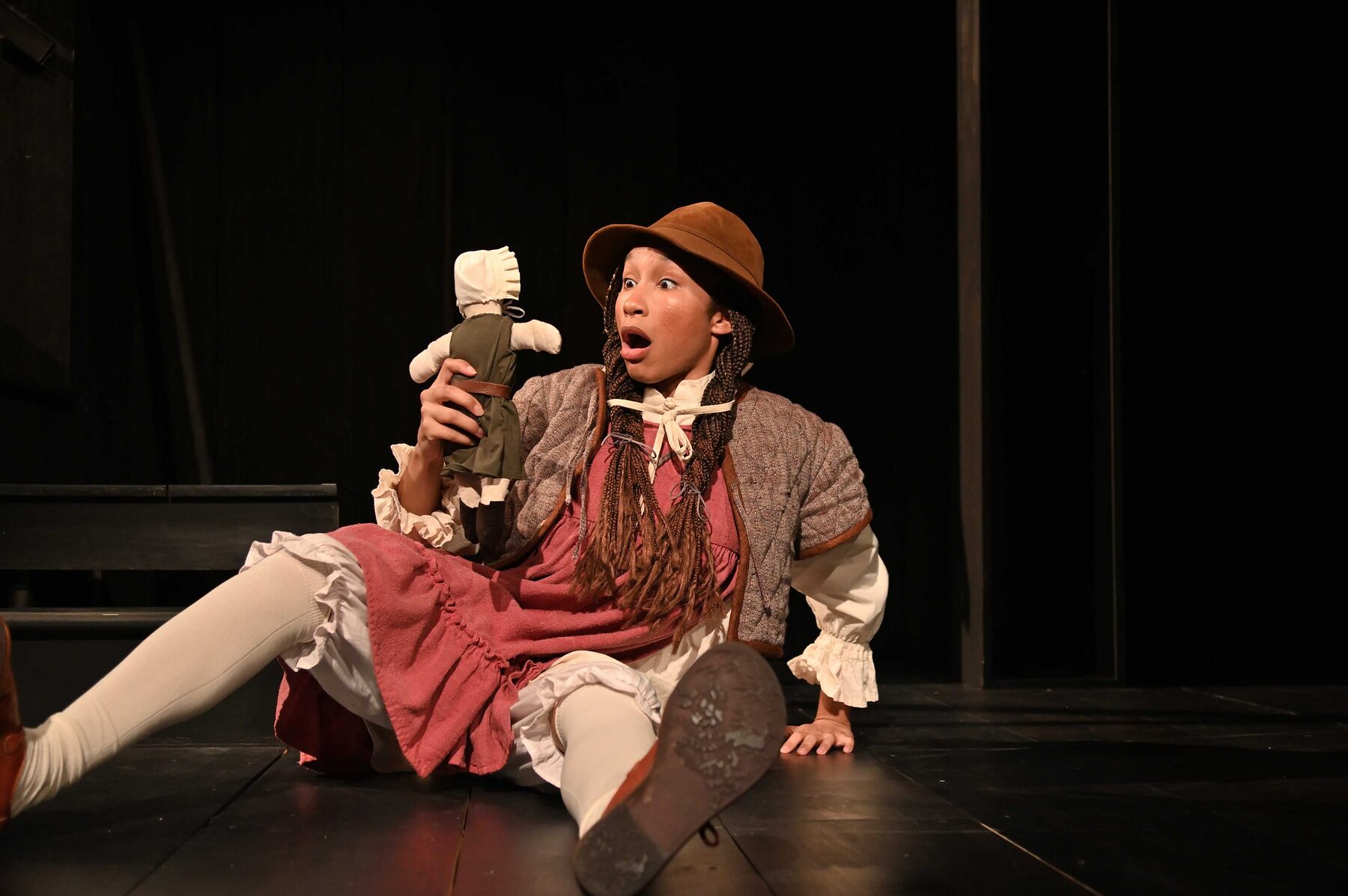 A young girl in a brimmed hat sits on the stage floor holding a cloth poppet