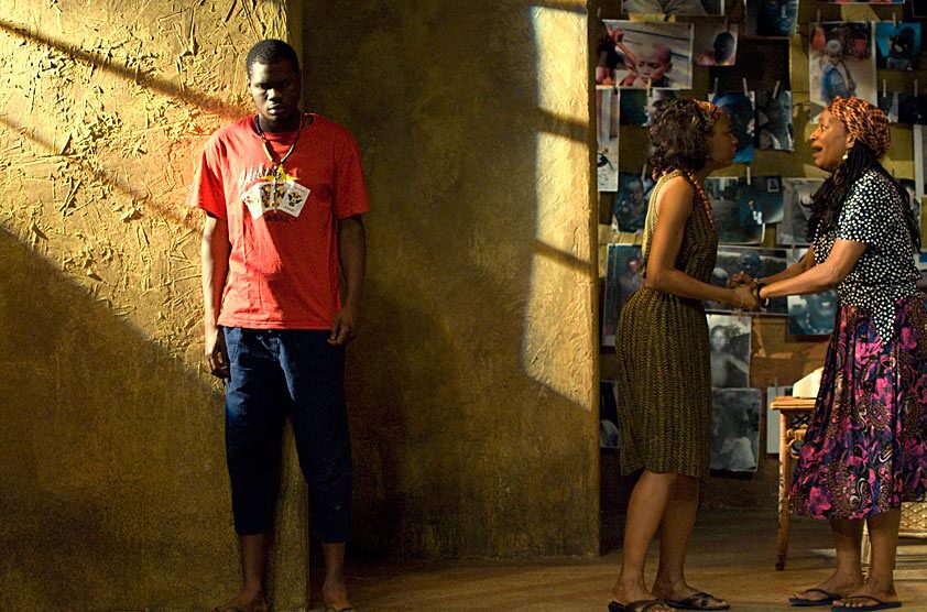 A young man stands apart from two women beside a wall of family photographs in The Overwhelming