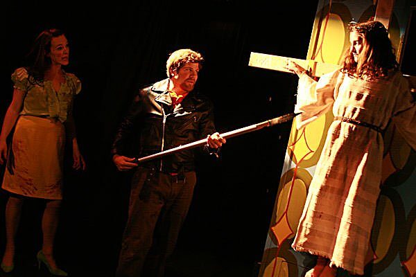 A man holding a wooden plank between two young women in The Pillowman