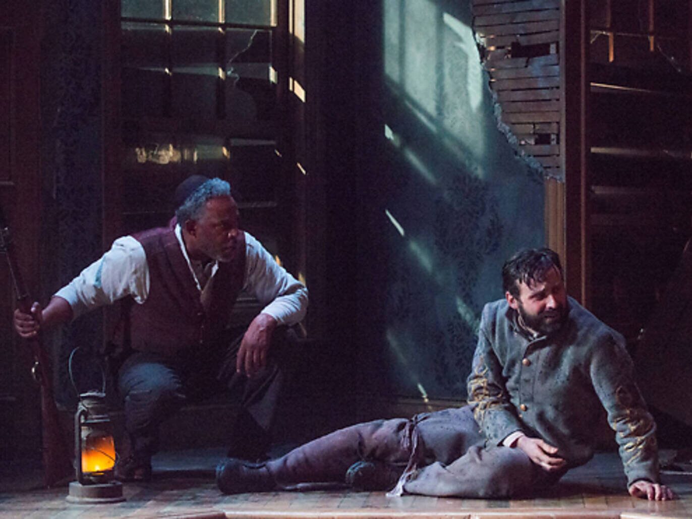 A wounded man on the floor with another kneeling over him by lantern light