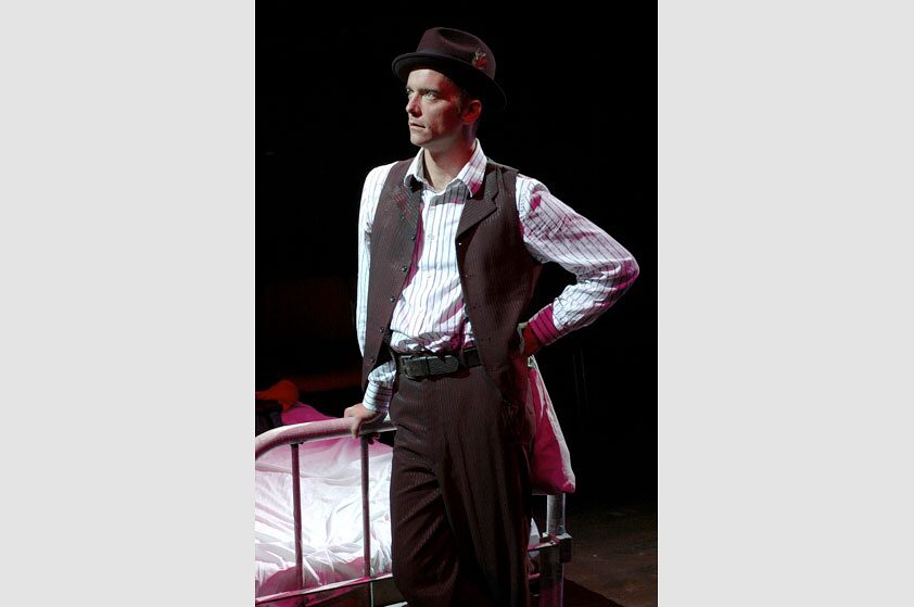 A man in a period vest and hat stands by an iron bedframe in Thieves Like Us