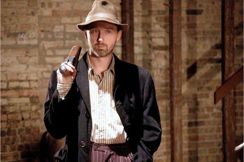 A man in a fedora holding a gun against a brick wall in Thieves Like Us